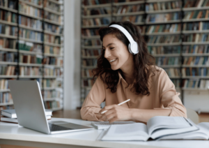 A student is in a library, watching the screen of a laptop, with a pen in their hands and a notebook on the table.